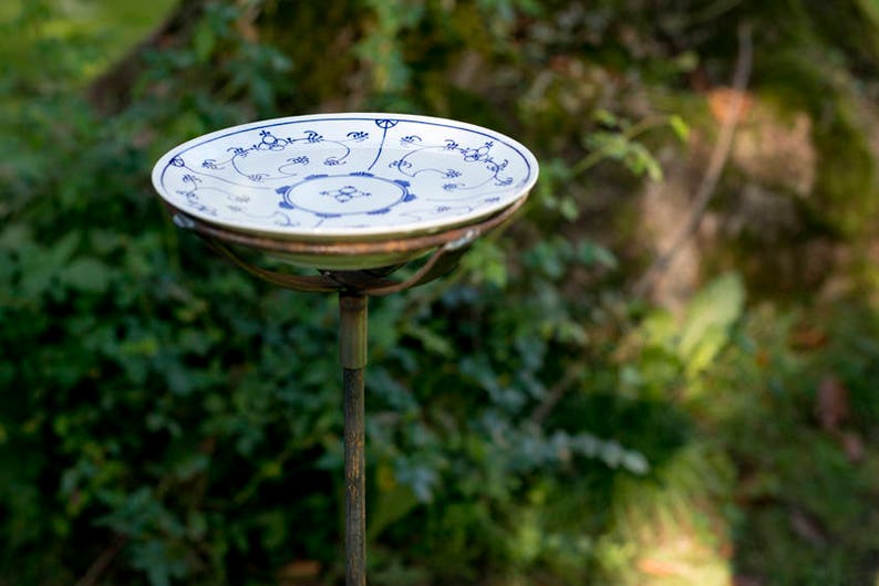 May include: A blue and white ceramic bird bath with a metal stand. The bird bath is on a metal stand with a round base. The bird bath is in a garden setting.