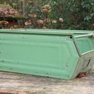 Könnte beinhalten: Eine verwitterte, grüne Metall-Aufbewahrungsbox mit rechteckiger Form und schräger Vorderseite. Sie hat einen Griff und steht auf einer Holzoberfläche. Die Farbe ist abgenutzt, was auf Alter hindeutet. Der Hintergrund ist unscharf, mit Blättern. Maße: 40cm x 20cm x 20cm.