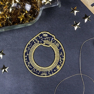 May include: A navy blue embroidered patch with a gold snake design. The snake is curled into a circle and has a variety of celestial symbols embroidered on it, including stars, moons, and suns.