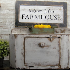 May include: White painted wooden cabinet with a single door and a shelf. The cabinet has a distressed finish and is decorated with a sign that reads "Welcome to our FARMHOUSE".