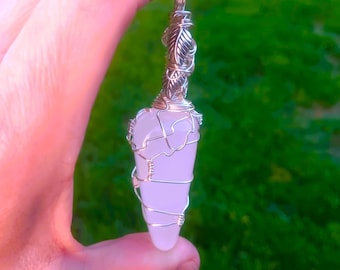 Puede incluir: Colgante de cristal rosa claro envuelto en alambre plateado. La parte superior del colgante presenta un diseño de hojas decorativas. El cristal en forma de lágrima se sujeta con el alambre.