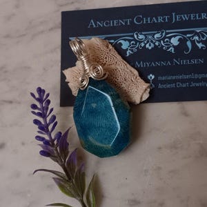 May include: A teal blue gemstone pendant with silver wire wrapping. The pendant is hanging from a silver chain.