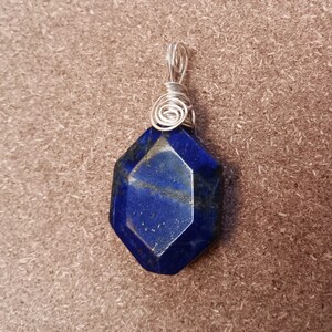 May include: A faceted, deep blue lapis lazuli pendant with silver wire wrapping. The gemstone is a hexagonal shape with visible gold flecks. The wire forms a spiral at the top for hanging.