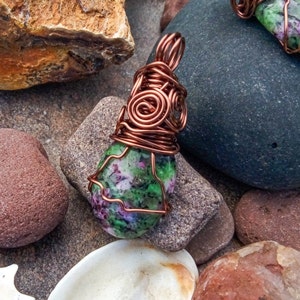 May include: A handcrafted pendant featuring a polished, oval-shaped stone with green and purple hues, wrapped in copper wire. The wire is intricately coiled at the top, creating a decorative bail for a necklace. The pendant rests on a bed of various stones.