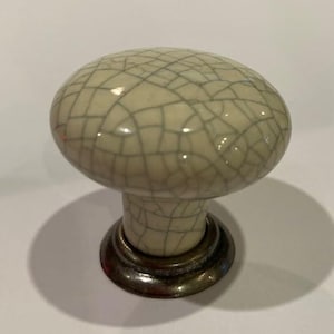 May include: A cream ceramic cabinet knob with a crackle glaze design. The knob has a rounded top and a dark metal base. This decorative hardware is suitable for drawers and cabinets.