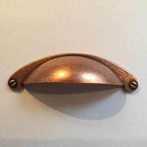 May include: A copper-toned cabinet pull with a rounded, arched shape. The pull has a slightly textured finish and is designed for use on drawers or cabinets.