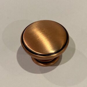 May include: A copper-coloured cabinet knob with a brushed metal finish. The knob has a rounded top and a cylindrical base, with a black accent ring. The hardware is designed for use on drawers or cabinet doors.