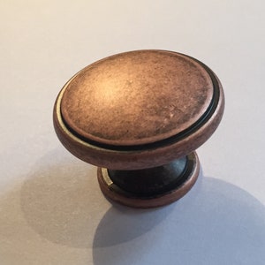 May include: A round, copper-coloured cabinet knob with a black ring around the edge. The knob has a textured surface and a black base.