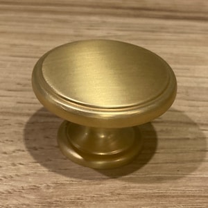 May include: A gold-toned cabinet knob with a round top and a slightly raised base. The knob has a smooth, brushed finish.