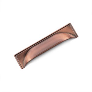May include: A copper-toned cabinet pull with a slightly curved rectangular shape. The pull has a smooth, brushed finish.