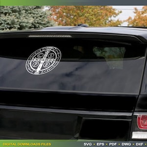 May include: A black car with a white Saint Benedict medal sticker on the back window. The medal features a cross with the letters 'C.S.S.M.L.  N.D.S.M.D.' around it.