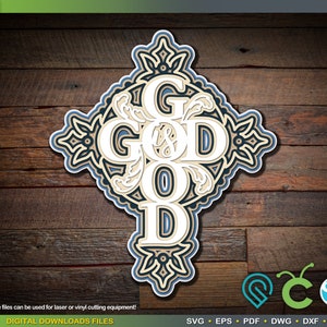 Könnte beinhalten: Ein weißes Kreuz mit dem Wort "GOD IS GOOD" in einer dekorativen Schriftart. Das Kreuz ist von einem blauen und schwarzen floralen Design umgeben. Der Hintergrund ist eine Holzmaserung.