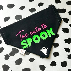 May include: Black dog bandana with the text "Too cute to spook" in pink and green lettering. A white ghost graphic is in the bottom right corner.