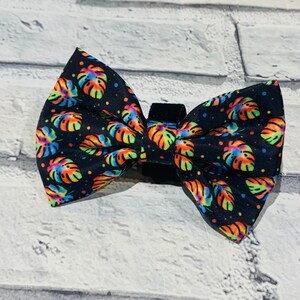 May include: A black bow tie featuring a colourful tropical leaf pattern. The leaves are in shades of orange, yellow, green, blue and pink. The bow tie has a black elastic band for attaching it to a collar.