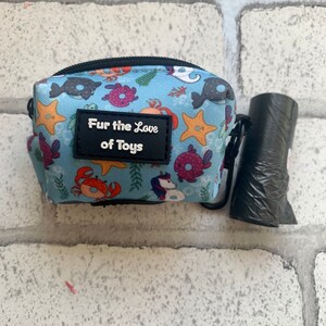 May include: A blue and white canvas bag with a zipper closure and a black strap. The bag features a variety of sea creatures printed on it, including crabs, starfish, and fish. The bag has a black patch with the text "Fur the Love of Toys" printed on it. The bag is sitting on a white brick surface next to a black plastic bag.