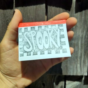 Può includere: Una scatola di fiammiferi rettangolare con un bordo superiore rosso brillante e un'etichetta bianca con la parola SPOOKY scritta in grandi lettere nere in grassetto. Il testo è circondato da un bordo a scacchi bianchi e neri su sfondo chiaro.