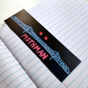 May include: A rectangular black bookmark featuring a light blue line drawing of a truss bridge spanning across the top. Below the bridge, the word MOTHMAN is printed in bold red capital letters, accompanied by two small, circular red dots.