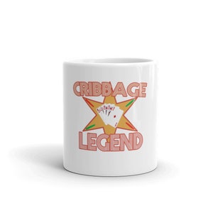 Pode incluir: Caneca de cer&acirc;mica branca com um design vermelho e laranja que diz "Cribbage Legend" com uma estrela amarela e cartas de jogo no centro.