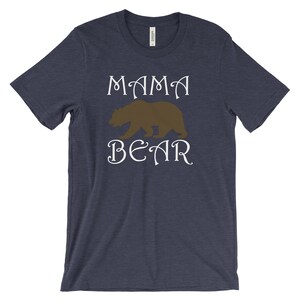 以下が含まれることがあります： 白い文字で「ママベア」と書かれたネイビーブルーのTシャツ。茶色の熊のシルエットが単語の間に中央に配置されています。