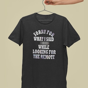 May include: A dark grey t-shirt with a white graphic that says "Sorry for what I said while looking for the remote" with a picture of a remote control.