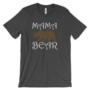 以下が含まれることがあります： 暗いヘザーグレーのTシャツに、白い文字で「ママベア」と書いてあります。文字の間に茶色の熊のシルエットが中央に配置されています。