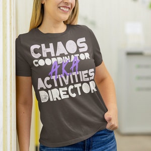 May include: A dark gray t-shirt with the text "Chaos Coordinator AKA Activities Director" in white with a distressed effect. The text "AKA" is in purple.