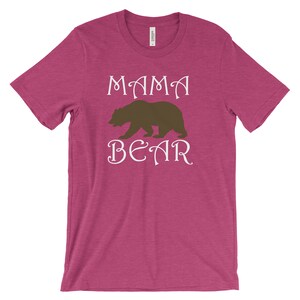 以下が含まれることがあります： 白い文字で「MAMA BEAR」と書かれたピンクのTシャツ。文字の間に茶色の熊のシルエットがあります。