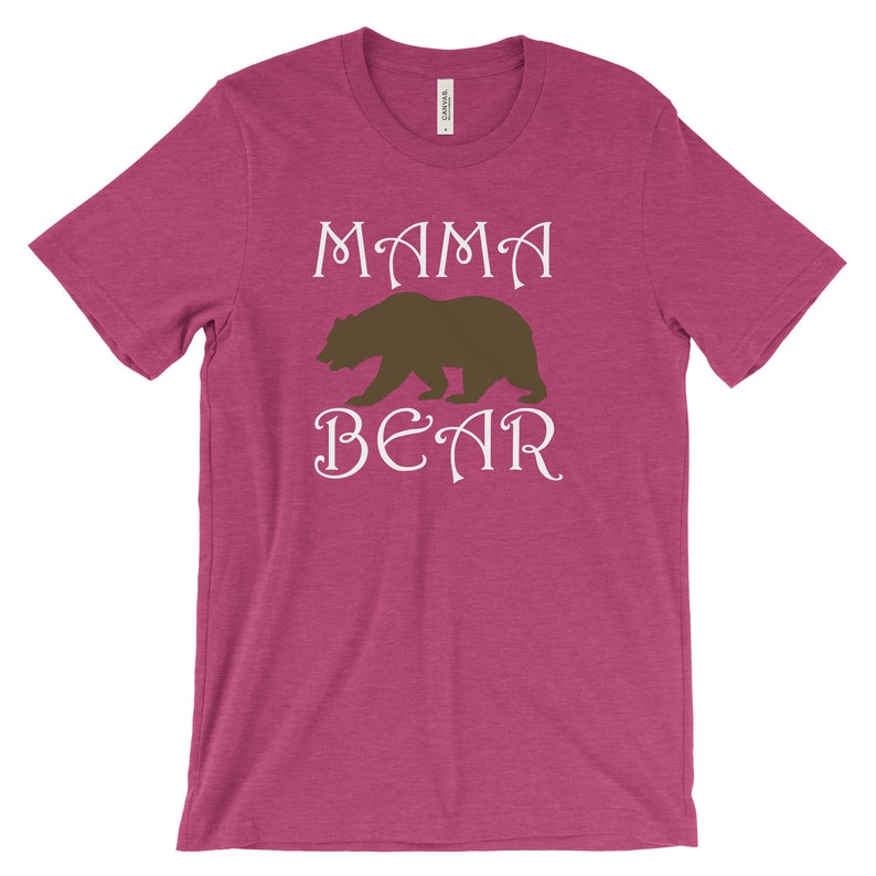 以下が含まれることがあります： 白い文字で「MAMA BEAR」と書かれたピンクのTシャツ。文字の間に茶色の熊のシルエットがあります。