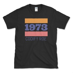 Peut inclure: T-shirt noir avec un motif graphique composé de bandes horizontales orange, rose et bleues. Le graphique comprend l'année "1973" et le texte "CODIFY ROE".