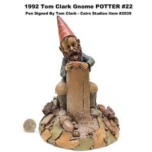 May include: A detailed figurine of a gnome potter, seated on a wooden post, wearing a red hat and holding a small bowl. The base features leaves and acorns. The text "1992 Tom Clark Gnome POTTER #22" is visible.
