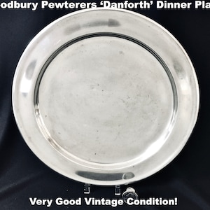 May include: A silver-colored 'Danforth' dinner plate by Woodbury Pewterers. The plate has a wide rim and a slightly concave center. The text "Very Good Vintage Condition!" is at the bottom. The plate is displayed on a clear stand against a black background.