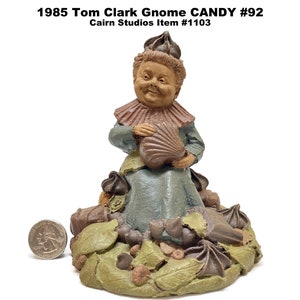 May include: A vintage Tom Clark Gnome Candy #92 figurine from 1985. The gnome, with a smiling face, wears a ruffled collar and holds a seashell. The base features leaves, nuts, and chocolate candies. The figurine is approximately 4 inches tall.