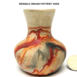 May include: A handmade Nemadji Indian pottery vase with a bulbous body and flared neck. The vase features a cream-colored base with swirling patterns of red, orange, and gray. The text "NEMADJI INDIAN POTTERY VASE" is at the top.