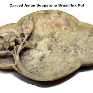 May include: A carved Asian soapstone brush/ink pot. The light-colored stone has a floral carving on one side. The pot is oval-shaped with a shallow basin and a reddish-brown hue on the edges.