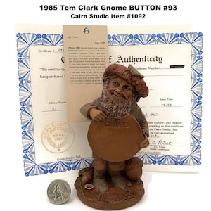 May include: A brown gnome figurine holding a round sign with the word "CAIRN" on it. The figurine is part of the 1985 Tom Clark Gnome BUTTON #93 collection. Includes a certificate of authenticity and a quarter.