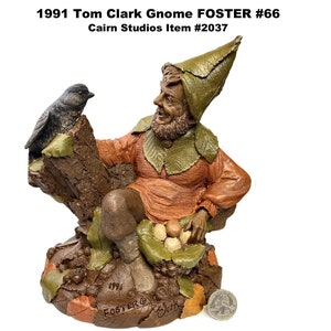 May include: A detailed figurine of a gnome, wearing a leaf-shaped hat and orange tunic, seated with a bird perched on a tree trunk. The base features leaves and nuts. Text on the figurine reads "1991 Tom Clark Gnome FOSTER #66".