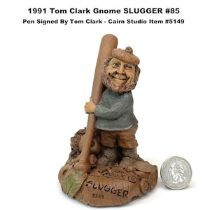 May include: A detailed figurine of a gnome holding a baseball bat, labeled "SLUGGER." The gnome wears a hat, sweater, and pants. The base is inscribed with the text "1991 Tom Clark Gnome SLUGGER #85 Pen Signed By Tom Clark - Cairn Studio Item #5149."