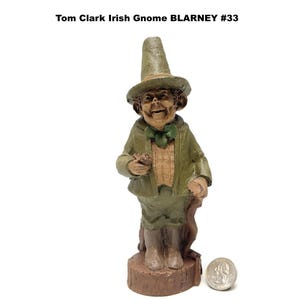 May include: A figurine of an Irish gnome named Blarney, wearing a green hat, jacket, and pants. The gnome is holding a small object and leaning on a wooden cane. The figurine is on a brown base, with the text "Tom Clark Irish Gnome BLARNEY #33" above it.