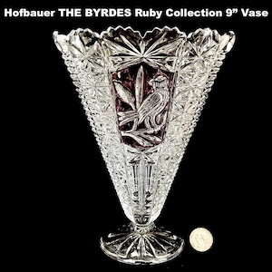 May include: A clear, cut-glass vase from the Hofbauer THE BYRDES Ruby Collection, approximately 9 inches tall. The vase features a triangular shape with a detailed bird and floral design in a ruby-colored panel. The vase has a starburst pattern and a circular base.