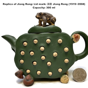 May include: A green ceramic teapot with a frog figurine on the lid. The teapot has a textured surface with small, round, light-colored accents. The text on the image reads "Replica of Jiang Rong: Lid mark: 蒋蓉 Jiang Rong (1919~2008) Capacity: 300 ml."