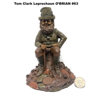 May include: A detailed figurine of a seated leprechaun, wearing a green hat and jacket, with a long beard and holding a walking stick. The base is adorned with leaves and a coin. The text "Tom Clark Leprechaun O'BRIAN #63" is at the top.
