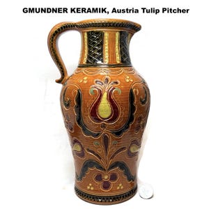 May include: A tall, brown ceramic pitcher with a handle, featuring a tulip design. The pitcher has a wide base and a narrow neck, with decorative floral patterns in red, yellow, and black. The text "GMUNDNER KERAMIK, Austria Tulip Pitcher" is at the top.