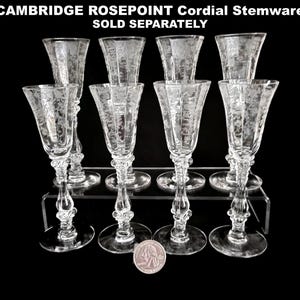 May include: Clear Cambridge Rosepoint cordial stemware glasses with etched floral designs. The glasses are displayed on a clear acrylic stand against a black background. The text "CAMBRIDGE ROSEPOINT Cordial Stemware SOLD SEPARATELY" is at the top.
