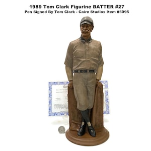 May include: A figurine of a baseball player in a vintage uniform, leaning against a wooden structure. The figurine is brown and tan, with a black belt and shoes. The text "1989 Tom Clark Figurine BATTER #27" is visible.