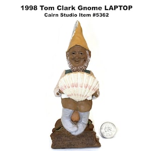 May include: A whimsical gnome figurine holding a seashell, wearing a tan hat and vest, and light blue pants. The figurine is on a brown base. The text "1998 Tom Clark Gnome LAPTOP" is at the top.