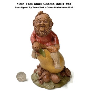 May include: A figurine of a smiling gnome, seated on a pear, with a beard and red shirt. The base features acorns and leaves. Text on top reads "1981 Tom Clark Gnome BART #41 Pen Signed By Tom Clark - Cairn Studio Item #134". A coin is next to the figurine.