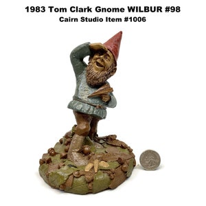 May include: A figurine of a gnome named Wilbur, from 1983, with a red hat and green tunic, holding a book. The gnome is standing on a base of leaves and acorns. Text on the image reads "1983 Tom Clark Gnome WILBUR #98" and "Cairn Studio Item #1006".