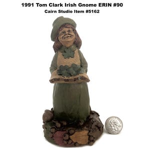 May include: A ceramic figurine of an Irish gnome, wearing a green hat and dress, holding a tray of shamrocks. The figurine has a smiling face and is standing on a base with leaves and a coin. The text on the tray reads "ERIN GO BRAGH".