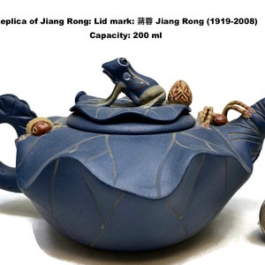 May include: A blue ceramic teapot with a frog figurine on the lid. The teapot is shaped like a lotus flower with leaf details. The text on the image reads "Replica of Jiang Rong: Lid mark: 蒋蓉 Jiang Rong (1919-2008) Capacity: 200 ml."