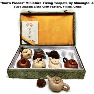 May include: A set of miniature Yixing teapots in a decorative box. The box is green with a gold interior and contains several small, uniquely shaped teapots in shades of brown, beige, and gray. The text "Sun's Pieces" is visible.